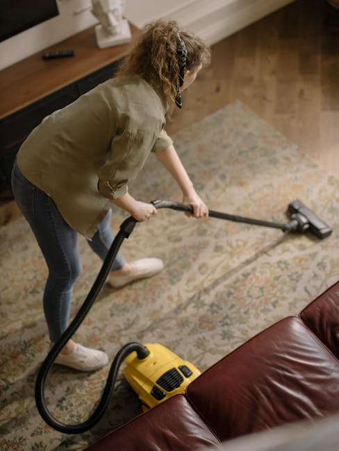 A woman using a yellow vacuum cleaner with a long hose attachment to deep clean a patterned area rug in a living room. She is leaning forward, focusing on the surface, which appears clean and well-mai